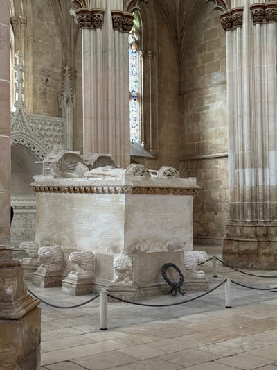 This image displays the interior of the Founder's Chapel within the Batalha Monastery (Mosteiro da Batalha) in Batalha, Portugal. The central subject is a large, rectangular stone tomb, specifically the joint tomb of King John I and Philippa of Lancaster. The tomb's base is supported by six carved stone lions. Its main body features carved inscriptions on its visible sides and an intricate frieze along the top edge. The lid presents two recumbent effigies, crowned and resting on carved pillows, partially obscured by ornate, shell-like carved canopies.

The surrounding environment includes several massive, light-colored, fluted stone columns with ornate capitals, supporting Gothic-style pointed arches and ribbed vaulting. Walls are constructed from cut stone blocks. A tall, narrow stained glass window with multicolored panes is visible in the mid-background. The floor is paved with large, rectangular stone tiles. A low barrier, composed of a dark chain strung between light-colored cylindrical bollards, restricts access to the tomb. A dark wreath rests on the tiled floor against the base of the tomb on the right side of the barrier. A portion of another ornate carved stone structure is visible in the far-left background. No individuals are present in the scene.