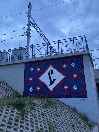 This vertical outdoor photograph, captured in Subotica, Serbia, features a vibrant mural painted on a concrete wall, likely beneath an overpass or bridge. The mural, resembling a traditional rug or tapestry, is predominantly blue with intricate red and white geometric patterns, including a serrated border. At its center, a large white diamond contains a distinct, stylized black number "7".
To the left, a tall, dark metal lattice structure, indicative of a power line tower or railway overhead line support, stands against an overcast, cloudy sky. Multiple thin wires stretch across the sky from this structure. The diffused light suggests a cloudy daytime, possibly late afternoon or early morning.
On the ground to the right of the mural, a sloped area is covered with gray concrete grid pavers, through which green grass and some dry weeds grow.
Visible on the concrete wall above the gridded ground, to the right of the mural, is black Cyrillic text. It reads: "ФОНДАЦИЈА ЗА СРПСКИ НАРОД И ДРЖАВУ," which translates to "Foundation for Serbian People and State." Below this text, faint markings resembling "V - V" can be discerned.