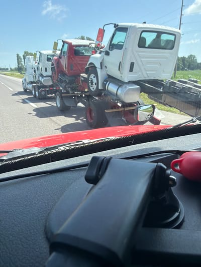 The image depicts a convoy of flatbed trucks carrying other trucks as cargo on a sunny day. The trucks are traveling on a paved road, likely a highway, with a shoulder and a grassy area with some trees to the right. The sky is blue with some scattered white clouds. In the foreground, the dashboard of a red vehicle is visible, suggesting the photo was taken from inside another truck. The trucks in transit appear to be new or recently manufactured, with some of them in white and others in red. The convoy is moving forward, as indicated by the motion blur on the road and the slight angle of the trucks. The scene is set in Hayti, Missouri, United States of America, as evidenced by the presence of power lines common in rural American landscapes.