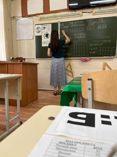 This image captures the interior of a classroom in Kaspiysk, Russia, focused on a female figure writing on a blackboard. The classroom features light-colored walls, a wooden floor, and artificial overhead lighting from two long fixtures above the blackboard. Natural light enters from a window on the left, partially obscured by vertical blinds.

On the left side of the room, a wooden teacher's desk and chair are visible. In the foreground, a light-colored student desk holds several papers. One paper prominently displays a large black Cyrillic letter "Я:" (Ya:). Another paper below it shows a table listing subjects like "Русский язык" (Russian language), "Математика" (Mathematics), "Физика" (Physics), and "Химия" (Chemistry) with corresponding codes "01", "02", "03", and "04". A dark coin rests on this desk.

The primary subject is a female individual, seen from behind, standing in front of a large green blackboard. She wears a dark green top, a long patterned skirt (grey and white tones), and dark sandals. Her long dark hair is down. She is actively writing on the blackboard with chalk in her right hand.

The blackboard is meticulously formatted with chalk-drawn boxes and text. Visible headings include: "КОД РЕГИОНА" (REGION CODE) with "15" entered, "КОД ОБРАЗОВ ОРГАНИЗАЦИИ" (EDUCATIONAL ORGANIZATION CODE), "КЛАСС БУКВА НОМЕР" (CLASS LETTER NUMBER) with "09" entered, and "КОД ППЭ" (P.P.E. Code, likely Exam Conducting Point Code) with "0481" entered. Below these, "НАЗВАНИЕ ПРЕДМЕТА" (SUBJECT NAME) has "МАТЕМ" (likely truncated "Математика" for Mathematics) written in a box. Further right, "ПОДПИСЬ СТРОГО В РАМКЕ" (SIGNATURE STRICTLY WITHIN THE FRAME) and "ДОКУМЕНТ СЕРИЯ НОМЕР" (DOCUMENT SERIES NUMBER) are visible. A small pink cloth hangs on the bottom edge of the blackboard.

Mounted on the wall to the left of the blackboard are three signs: a "ВНИМАНИЕ!" (ATTENTION!) sign stating "Ведётся аудио и видео запись" (Audio and video recording is taking place), a "no phones" icon, and a "no talking" icon, below which are the numbers "0025". A flat-screen television is mounted high above the blackboard, and a long, horizontal heater or radiator is affixed to the wall below the TV. In the midground, various student desks and chairs are arranged, with one desk covered by a green tablecloth. A wooden set square is leaning against the wall on the right.