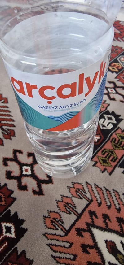 A close-up, high-angle shot shows a partially filled plastic bottle of "arçalyk" brand still drinking water. The bottle is sitting on a patterned rug with a predominantly beige background and geometric designs in red, black, and white. The label on the bottle features the brand name in large, orange, stylized letters, with "GAZSYZ AGYZ SUWY" and "STILL DRINKING WATER" printed in smaller blue text below. The water inside the bottle is clear, and condensation is visible on the outside of the plastic. The bottle's ribbed texture is evident. The scene appears to be indoors, possibly in a residential setting, given the presence of the rug. No people are visible. The overall impression is a still life of a beverage container.