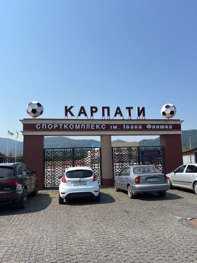 This is a full-frame, eye-level, outdoor, daytime, vertical photograph of the entrance to a sports complex in Khust, Ukraine. The entrance is a brick archway with a large sign above it. The sign has the word "КАРПАТИ" in large, red, capitalized letters, with two large soccer balls, one on each side, placed on top of the arch. Below this, another sign reads "СПОРТКОМПЛЕКС ім. Івана Фляшка".
In front of the archway, a black SUV is parked on the left, and a white hatchback is parked in the center. To the right of the white hatchback, a silver sedan is parked. The ground in front of the entrance is paved with cobblestones. Behind the entrance, a metal gate is visible, and beyond that, rolling hills and trees under a clear blue sky. Two Ukrainian flags are mounted on flagpoles to the left of the entrance. A smaller sign with Cyrillic text and graphics is affixed to the right side of the entrance arch.