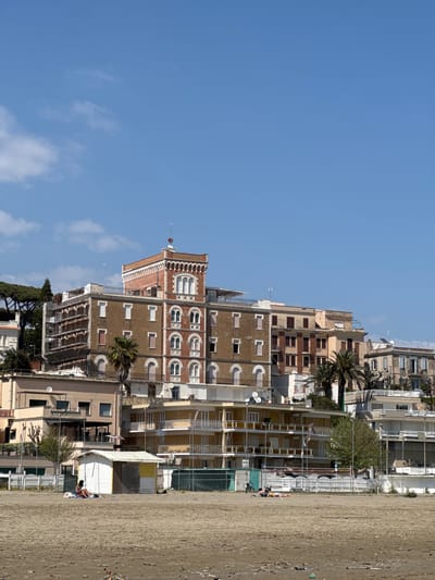 The image shows a vertical view of buildings and a beach in Anzio, Italy, under a bright blue sky. A prominent brownish building with arched windows and a tower stands out. Other buildings of varying heights and architectural styles are clustered together. A sandy beach occupies the right side of the frame, with a couple of people visible. Palm trees are interspersed among the buildings, suggesting a coastal environment.