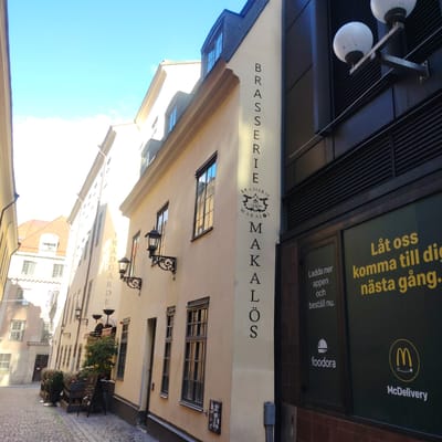 The image displays a narrow, sunlit cobbled street in Stockholm, Sweden, specifically Trädgårdsgatan, identifiable by the vertical "TRÄDGÅRDE" text on the left building and "BRASSERIE MAKALÖS" on the central building. The scene is dominated by architectural elements under a clear, bright blue sky.

On the left, a light-colored, multi-story building with numerous windows and a visible red-tiled roof in the background lines the street. The vertical text "TRÄDGÅRDE" (part of "Trädgårdsgatan") is visible on its facade.

The central building, Brasserie Makalös, features a light beige facade with multiple rectangular windows and three black, ornate wall-mounted lamps. The name "BRASSERIE" is vertically written in black capital letters above a decorative logo, and "MAKALÖS" is written similarly below the logo. An outdoor seating area with wooden planters containing green foliage and a black A-frame sign is positioned on the cobblestone pavement in front of this building.

To the far right, an adjacent building presents a modern, dark facade with horizontal and vertical slatting. Two white, spherical wall lamps are mounted on its upper section. A large window or display on the lower part of this building features advertisements in yellow and white text against a dark green background. The yellow text reads "Låt oss komma till dig nästa gång." and the white text below says "Ladda ner appen och beställ nu." Prominently displayed are the white "foodora" logo (a chef's hat over a plate) and the yellow "McDelivery" logo (McDonald's "M" with a delivery circle).

The cobblestone street appears clean and empty of people or vehicles. Strong sunlight illuminates the scene, casting distinct shadows. No individuals, actions, or direct interactions are visible.
