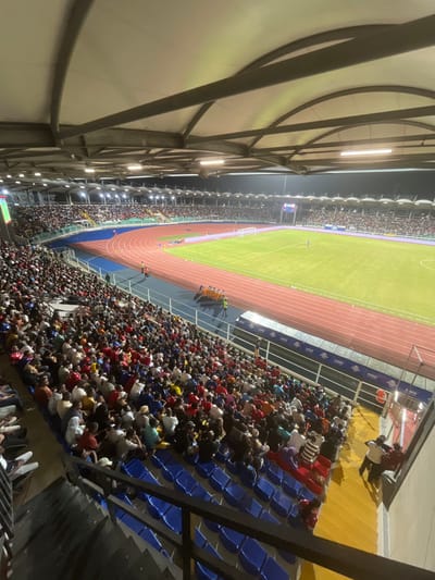 A stadium packed with spectators. A running track surrounds a green field. Stadium lights illuminate the crowd. The seating area is filled with people, many wearing hats and colorful shirts. The seats are primarily blue. A structure with white walls and metal supports flanks the stadium.
