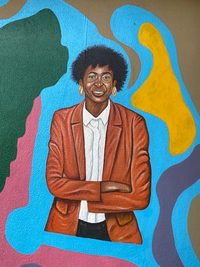 The image is a close-up of a mural. It depicts a brown-skinned woman with dark hair and gold earrings, wearing a white shirt and a reddish-brown blazer. She is smiling and has her arms crossed. The background consists of abstract shapes painted in various colors, including blue, yellow, green, pink, and purple. The image does not contain enough information to determine if it is in the city of Tawpo, Nigeria.