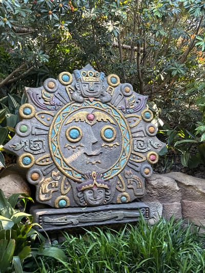 A carved stone sun disk artifact with intricate, colorful designs, rests on a stone pedestal in a lush garden. The artifact is gray with accents of gold, turquoise, and pink, depicting a central face surrounded by decorative motifs. Behind the artifact, lush green foliage and trees with small fruits are visible. The scene is well-lit, suggesting it is daytime. This artifact is located in Pudong, China, likely within a botanical garden or themed attraction.