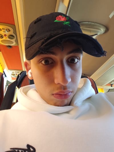 A young man takes a selfie on what seems to be a bus. He is wearing a black baseball cap with a red rose embroidered on it, a white hoodie, and a single white wireless earbud in his left ear. The bus interior includes orange panels and a ceiling light. His expression is earnest, with wide eyes.