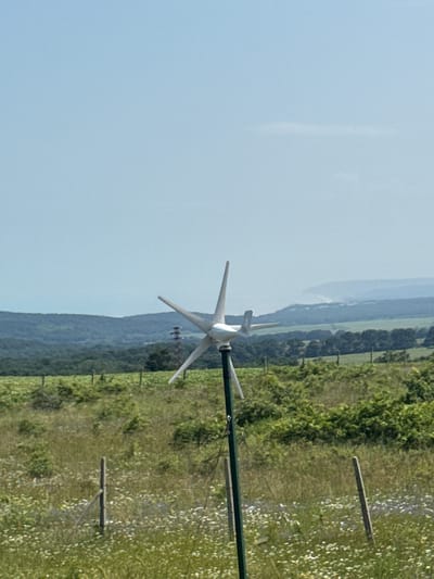 The image captures a rural landscape in what appears to be Bliznatsi, Bulgaria, under a bright, hazy blue sky. In the foreground, positioned towards the right, stands a small, white, three-bladed wind turbine, mounted on a tall, slender green pole. Its blades are visible and appear static or slowly turning.
The ground around the turbine is covered in lush green vegetation, resembling a field of grass possibly interspersed with wildflowers, which appear as scattered white and lighter green patches. Several rustic wooden posts are visible within this field, suggesting a fence line or marking.
Behind the immediate foreground, a dense line of trees or forest stretches horizontally across the mid-ground, forming a natural barrier. Beyond this treeline, distant rolling hills or mountains are visible, their forms softened and lightened by atmospheric haze, blending into the pale sky.
The scene conveys a peaceful, open, and natural outdoor environment, likely during daytime given the bright illumination. There are no people visible, and no discernible text is present. The focus is on the interplay of nature and a small-scale renewable energy structure.