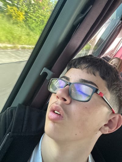 A person with short dark hair and wearing glasses is sleeping in a vehicle. The person is wearing a black jacket over a white shirt. The vehicle is moving, as evidenced by the blurred scenery visible through the window. Another person's face is visible in the background. The location is suggested to be in the Municipal Unit of Kato Olympos, Greece, but there is no visual information in the image to confirm this.