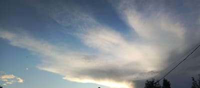 The image depicts a vast expanse of the sky above Merchalovo, Russia. The sky is a gradient of blue, transitioning from a lighter shade at the bottom left to a deeper hue towards the top. Wispy, white cirrus clouds stretch across the frame, appearing feathery and delicate. In the center right, a large, dark cumulonimbus cloud dominates the scene, with a distinct shelf-like formation at its base, suggesting the possibility of precipitation. The lower portion of the image reveals a dark silhouette of trees and a power line stretching diagonally across the right side. A faint glow of sunlight is visible behind the cumulonimbus cloud, indicating either sunrise or sunset. The overall impression is one of an approaching storm in a rural or semi-rural setting.