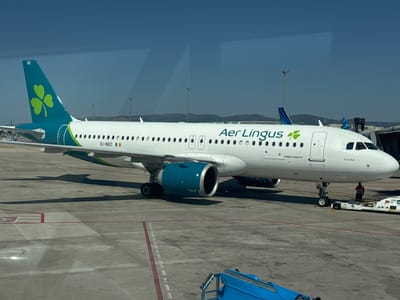 This media file shows an Aer Lingus Airbus A320 aircraft parked on a tarmac at an airport. The aircraft is predominantly white with a distinctive teal and green livery on its tail, featuring a prominent shamrock logo. The text "Aer Lingus" is visible on the fuselage, along with the aircraft model "AIRBUS A320 NEO" and its registration "EI-NSC". The name "Fearghal" is also visible on the nose of the aircraft.
The scene is set in what appears to be a sunny day, indicated by the clear blue sky and the shadows cast on the tarmac. The ground is concrete, marked with yellow and red lines typical of an airport runway or taxiway. In the background, other aircraft, airport structures, and distant hills can be seen. A ground crew member in a high-visibility vest is standing near the front landing gear, overseeing some equipment. A tow vehicle is attached to the front of the aircraft. The overall impression is of an aircraft preparing for or having just completed a flight. The location is identified as el Prat de Llobregat, Spain, which is the location of Barcelona–el Prat Airport.