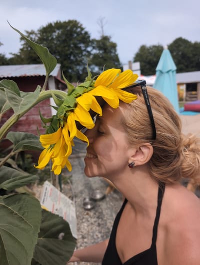 A blonde woman with her eyes closed and smiling is smelling a sunflower outdoors. She is wearing sunglasses on her head and a black halter top. The sunflower's yellow petals droop down, with some touching her forehead. The background shows trees, a building, and blue umbrellas. The scene appears to be in Saint-Jouin-Bruneval, France, suggesting a park or garden setting.