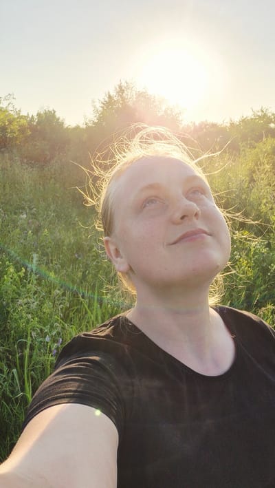 A young woman with fair skin, freckles, and blonde hair is looking up at the sun, which is setting behind trees in the background. She is wearing a black t-shirt. The scene is outdoors, with tall green grass and wildflowers in the foreground. The sun's rays create a golden light that illuminates her hair and face. The image appears to have been taken in Piatra Albă, Moldova.