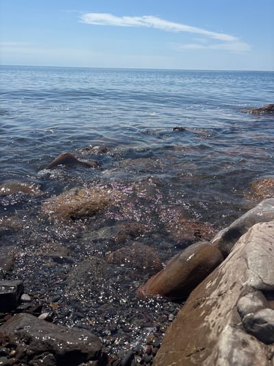 The image shows a rocky shoreline meeting the sea. Clear water laps against the rocks, with light reflecting off the surface. A few clouds are visible in the blue sky. No people are visible. The specific location in Semidvorye, Ukraine cannot be determined from the image alone.