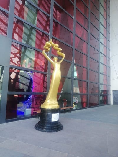 A large, golden, stylized statue of a female figure stands on a dark, multi-tiered circular base with a visible white plaque, positioned on a grey paved surface in Fengtai District, China. The figure, depicted in a flowing gown, raises its arms to hold a swirling, ribbon-like element above its head. Behind the statue, a modern building facade rises, composed of dark-framed, predominantly red-tinted glass panels. These panels exhibit strong reflections of internal elements, including blue and purple neon lighting, circular patterns, and sections of green-tinted glass. A wooden bench is partially visible against the facade to the right of the statue. To the far right, the building transitions to a lighter, solid wall section against which a dark sheet is draped. No individuals are present in the immediate foreground.