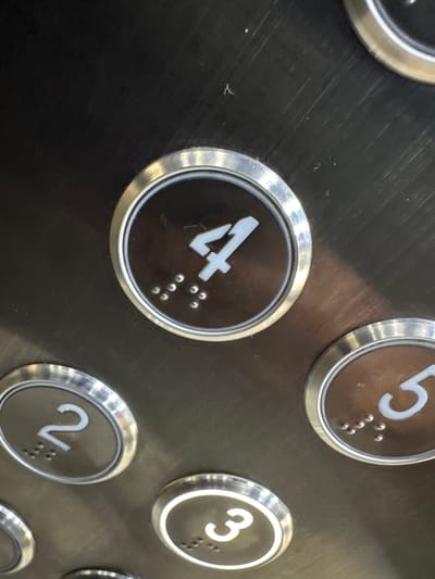 The image shows a close-up view of an elevator control panel, likely in Koltsovo, Russia. The panel is made of brushed metal and features several circular buttons with illuminated numbers and braille. The visible buttons include "4," "2," and "5," and a partial view of a button labeled "3" is also present. Each button is surrounded by a silver-colored trim. The button for the number "4" is centrally located and is the most prominent. It displays a large white numeral "4" and a series of braille dots below it. To the left of the "4" button is a button for "2," and to the right is a button for "5." The "3" button is visible towards the bottom of the frame. The overall environment appears to be indoors, within an elevator car. There are no people visible in the image. The metal surface of the panel shows some minor scuff marks and dust.