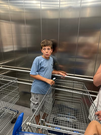 The image shows a young boy in a blue t-shirt and gray pants standing next to a metal shopping cart. The boy is looking at the camera with a slightly confused expression. His right hand is on the shopping cart, and he is holding a small, colorful object in his left hand. The shopping cart is in front of a reflective, metallic wall, possibly an elevator or a stall in a store.
Behind the boy, another person's legs are visible, wearing a white towel or cloth. Further to the right, another shopping cart with a blue handle is visible, with the IKEA logo and "www.ikea.com" printed on it. The overall scene appears to be indoors, possibly in a shopping center or a large retail store like IKEA. The lighting suggests it's daytime. The boy's pants have a graphic design with text that reads "MAKE YOUR MIND THINK THE BEST OF COOL".