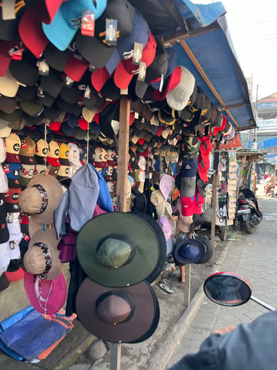 The image shows a street market in Gedang, Indonesia, likely selling hats. A large assortment of hats, including baseball caps, bucket hats, and wide-brimmed hats, are displayed on stalls and hangers. The hats are of various colors and styles, with some featuring logos and designs. The market appears to be outdoors, with a blue tarp overhead providing shade. In the background, a street with a few motorbikes and people can be seen. The scene conveys a sense of local commerce and everyday life in a Southeast Asian city.