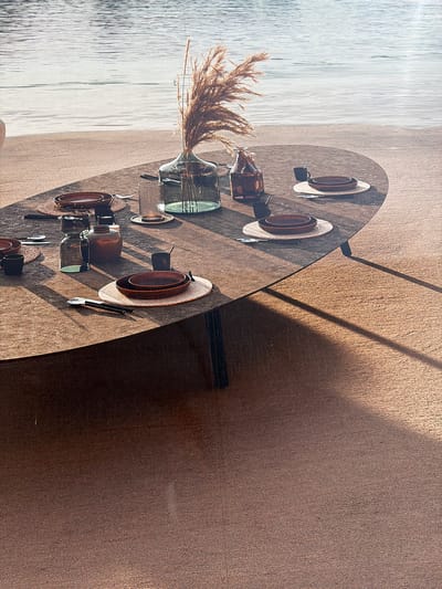 The image depicts an outdoor dining setting next to a body of water, likely a river or lake, given the gentle ripples and the sandy shoreline. The scene is situated in Mol, Belgium. A large, oval-shaped table, made of a textured, cork-like material, is set for a meal. The table has a warm, earthy tone.
On the table, there are several place settings. Each setting includes a round, brown placemat, topped with a larger, shallow brown bowl and a smaller, deeper brown bowl nested within it. Cutlery, consisting of a knife and fork, is placed on the left side of each setting. There are also small, dark mugs or cups at some settings.
A prominent feature of the table is a large, clear glass vase filled with dried, feathery grasses, adding a natural, rustic element to the arrangement. Beside the vase, there are two dark brown glass bottles, one taller than the other. Another smaller, opaque brown vase or jar is also present on the table. A textured, clear glass tumbler sits on a coaster.
The table is positioned on a surface that appears to be made of the same cork-like material as the tabletop. Sunlight casts strong shadows across the table and the surrounding surface, indicating a bright, sunny day. The background shows the expanse of the water and the sandy bank. No people are visible in the image.