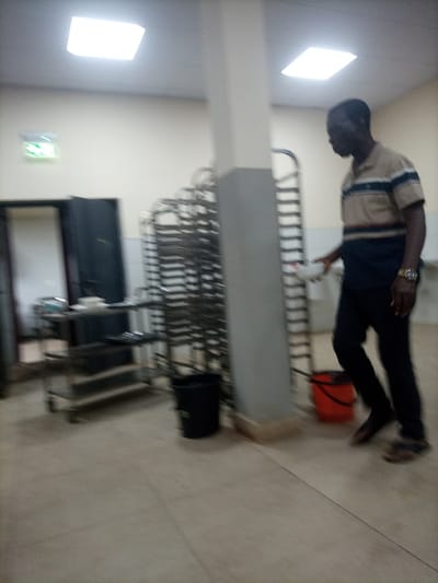 A man stands in a commercial kitchen. He wears a striped shirt, dark pants, and sandals, holding a white bowl. Stainless steel baking racks line a wall next to a gray pillar. A rolling cart with supplies and trays sits near an open doorway. Two trash cans, one black and one orange, are positioned on the tiled floor. Overhead, square fluorescent lights illuminate the room. A green exit sign glows above the doorway. The image is somewhat blurry, indicating movement.