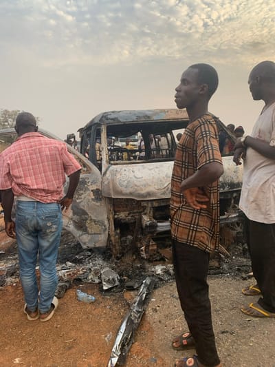 A burnt-out vehicle, possibly a bus, is surrounded by onlookers. Three men are in the foreground: one in a white shirt, another in a brown and black checkered shirt, and the third in a red plaid shirt and jeans. The vehicle's exterior is heavily charred, with visible interior details and potential passengers. Debris litters the ground around the vehicle. The sky is overcast, suggesting a somber or serious atmosphere.
