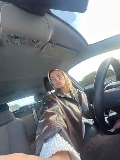 A female individual with medium-length blonde hair and clear-framed eyeglasses is seated in the driver's position of a passenger vehicle, taking a selfie. She is wearing a brown, leather-look jacket with visible white sherpa lining at the cuffs and collar, over a white inner layer. Her seatbelt is fastened across her chest, and her lips are pursed. The vehicle's interior features a light grey headliner, an overhead console with interior lights and a sun visor. A panoramic sunroof is visible above. A black steering wheel is partially visible on the right side of the frame. The background through the windows shows a bright outdoor setting with trees and distant vegetation, indicative of a road or highway environment. The scene is situated in Villeneuve-Loubet, France.