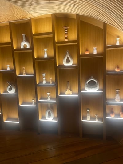 The image depicts an interior space, possibly a museum or gallery, featuring an arrangement of decorative ceramic objects displayed on wooden shelves. The shelves are illuminated from below, creating a warm and inviting ambiance. The objects themselves are varied in shape and size, with some appearing to be vases and others more abstract sculptures. The architectural style of the space, with its curved wooden ceiling, suggests a contemporary design. The presence of lighting fixtures and the careful presentation of the objects indicate that this is a curated exhibition or display. The scene is devoid of people, focusing entirely on the artistic arrangement of the ceramics within the architectural setting.
