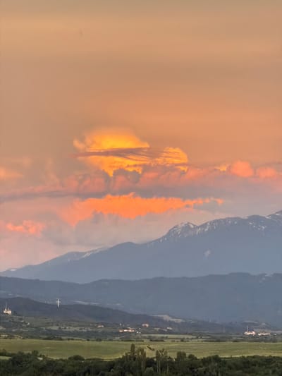 The image captures a serene landscape view from Kocherinovo, Bulgaria, at dusk. The sky is painted with hues of orange and pink, transitioning to a soft beige at the top, indicating the setting sun. Clouds, illuminated by the last rays of sunlight, create a dramatic visual spectacle in the upper part of the frame. Below the sky, layers of mountains dominate the middle ground. The closest mountain range is a dark, imposing silhouette against the vibrant sky, with patches of snow visible on its peaks. Further in the distance, additional mountain ranges fade into a soft blue haze.
In the lower portion of the image, the landscape transitions to rolling hills covered in lush green fields. A few scattered houses and buildings are visible, hinting at the presence of a settlement or village. Most notably, a large, white cross stands prominently on one of the hills, a significant landmark. To the left, a few more buildings, possibly part of a monastery or a church complex, are discernible. The foreground is a dense canopy of dark green trees, framing the entire scene. The overall atmosphere is tranquil and picturesque, highlighting the natural beauty of the Bulgarian countryside.