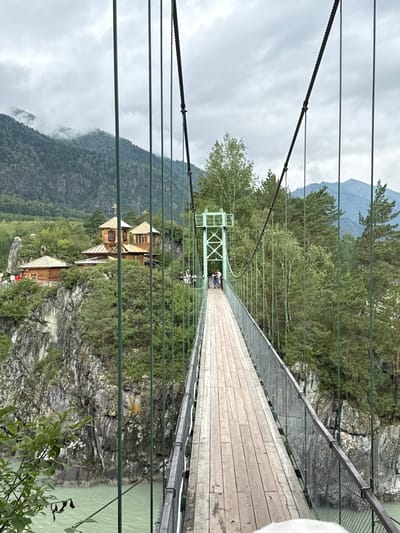 The image shows a suspension bridge in Chemal, Russia, crossing a body of turquoise water. The bridge has a wooden plank walkway with metal railings and is supported by cables. On the left side of the bridge, a cliff face overlooks the water, topped with a cluster of wooden buildings, including what appears to be a church with multiple golden domes and crosses. The surrounding environment is mountainous and forested, with lush green trees and a cloudy sky. A few people are visible on the bridge and near the buildings, suggesting a tourist location. The overall scene is one of natural beauty combined with man-made structures.