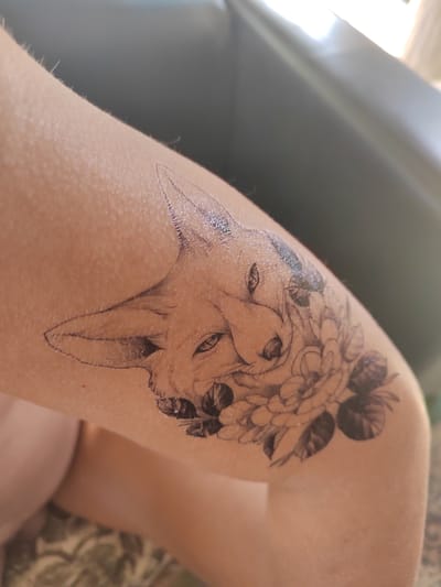 A close-up shot shows a temporary tattoo of a fox's head intertwined with flowers on a person's arm. The tattoo is rendered in black outline and shading, with the fox's face prominently displayed, its ears perked and eyes looking forward. Flowers, possibly roses and leaves, are depicted around the fox's head, adding a decorative element to the design. The skin appears fair and slightly textured, with fine hairs visible. The background is out of focus, suggesting an indoor or shaded outdoor environment.
