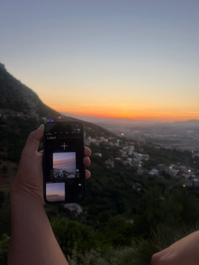 A panoramic shot captures a vibrant sunset or sunrise over a mountainous landscape, likely in Tétouan, Morocco, as indicated by the context. The sky transitions from a soft light blue on the left to a warm, glowing orange and yellow on the right, where the sun is low on the horizon. Below the horizon, a sprawling settlement with numerous buildings and dense green foliage covers the hills and valleys. Distant city lights appear as blurred bokeh against the darkening terrain.

In the lower right portion of the image, a person's arm and hand hold a black smartphone horizontally. The phone's screen is illuminated, displaying what appears to be a photo gallery or camera application interface. Visible on the screen are thumbnails of images, some of which depict sunset scenes similar to the actual view. Text elements on the phone's interface, extracted by OCR, include "Local Vault," navigation tabs like "MY," "MEU," "CORE," "TOP," "PPL," "NEAR," and categories such as "Latest," "Created," and "Investec." A large plus sign symbol ("+") is central on the screen, likely for adding new content, alongside a timestamp "05/31/26 20:30." The time of day is clearly dusk or dawn, with the warm hues of the sky dominating the scene. The image is taken from an elevated perspective, offering an expansive view of the city and nature.