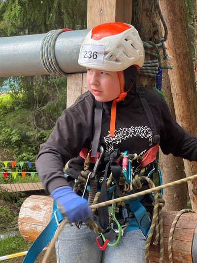 The image shows a person wearing a helmet and harness, suspended by ropes in what appears to be an outdoor adventure or obstacle course. The subject is wearing a white helmet with orange accents and a bib with the number "236" visible. They are also wearing blue gloves and a black hoodie with a white graphic design. The scene is set in a wooded area with trees and foliage visible in the background. Several ropes and carabiners are attached to the harness, indicating preparation for or participation in an activity like rock climbing or zip-lining. There are wooden structures, likely part of the course, and a series of colorful triangular flags in the background. The lighting suggests it might be daytime. The subject's expression is focused. The location context provided is Сергино, Russia.
