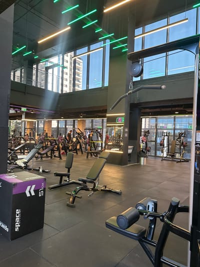 The image displays the interior of a modern, spacious fitness center, identified as being in Beer Yaaqov, Israel. The facility features a dark, rubberized floor and a high ceiling with exposed ductwork and numerous linear LED light fixtures, some emitting green and others warm white illumination. Large, floor-to-ceiling windows span the upper and lower levels, allowing natural light to enter and reflecting exterior multi-story buildings.

In the foreground, a black plyometric box with purple accents and "SPACE 51 CM" branding is visible on the left. Centrally, two black adjustable weight benches with dark green padding are positioned on the floor. On the far right, a portion of a black exercise machine with padded rollers is partially visible.

The mid-ground and background are populated with various exercise equipment, including multiple weight benches, barbell racks laden with purple and black weight plates, and diverse weight machines such as chest presses and leg presses. Approximately eight to ten individuals are present, distributed across the gym floor. Several are actively using equipment, while others are standing or moving between stations. A large black structural column stands prominently on the right, from which a lat pulldown cable system extends. Illuminated green and pink square signs are visible on the far walls, and a silver trash can is situated near a column.