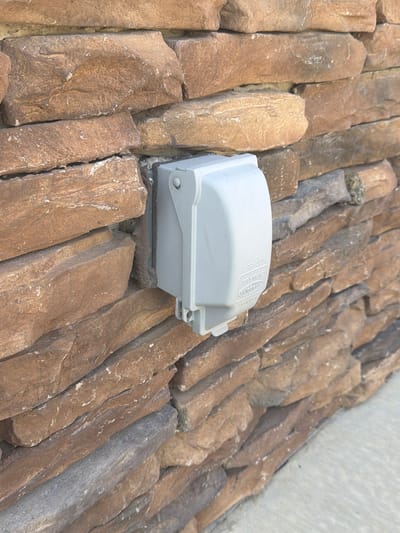 A light gray, rectangular electrical outlet cover is mounted on a stone wall. The stone is a mix of brown and gray hues, with varied shapes and sizes that create a textured, layered appearance. The cover has a circular button or latch visible on its top left. Some text is embossed on the cover, including "EXTRA-DUTY" and "TAYMAC". The outlet cover appears to be facing towards the viewer. The image is an outdoor, eye-level shot, with a shallow depth of field that blurs the background stones slightly. There are no people or animals visible in the image. The context of Cardwell, United States is unknown from the image alone.
