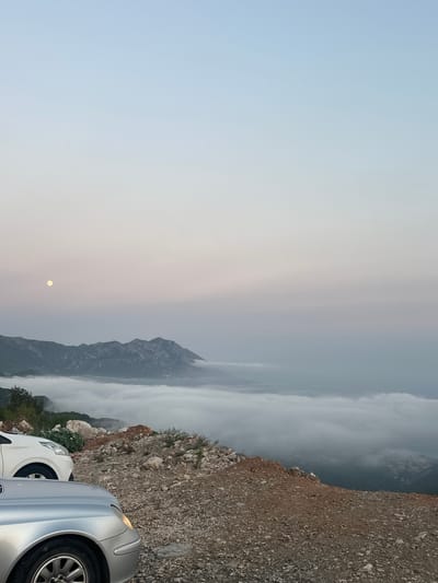 The image captures a serene mountainous landscape above a sea of clouds, likely taken from an elevated viewpoint near Lapčići, Montenegro. In the foreground, two cars are parked on a rocky, unpaved surface. The car on the left is a white SUV, with its front visible. Beside it, the front of a silver sedan is prominently featured, its headlight illuminated. Beyond the cars, the terrain slopes downwards, covered in loose stones and sparse vegetation.

The middle ground is dominated by a thick blanket of white clouds that obscures the valley below, creating an ethereal, dreamlike effect. Emerging from the clouds are the rugged peaks of mountains, their rocky textures and steep slopes clearly defined. In the upper left quadrant of the image, a pale full moon hangs in the soft, gradient sky. The sky transitions from a pale blue at the top to a muted pink and gray near the horizon, suggesting either dawn or dusk. The overall atmosphere is one of tranquility and majestic natural beauty.