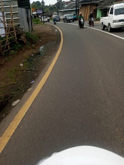 The scene depicts a curved asphalt road in the city of Chhenangri_Rishor, Bhutan, under an overcast sky. The image is captured from a low perspective, likely from a moving vehicle, with a white curved object visible at the bottom foreground.
The road features a solid yellow line delineating its left edge and a dashed white line separating lanes further to the right. The asphalt surface appears dark and partially damp or reflective. Along the immediate left shoulder, dirt is visible interspersed with sparse green vegetation, small puddles of water, and scattered litter including plastic debris.
Further left, a makeshift structure constructed from corrugated metal sheets and wooden scaffolding stands. Behind this, lush green trees and foliage extend up a gentle slope. In the mid-ground, a utility pole with multiple wires is present, along with a yellow road sign indicating a two-way traffic symbol. Partially visible beyond are several buildings with dark roofs and light-colored walls, consistent with urban architecture. One building exhibits a sign with the partial text "WARUNG."
On the road, traffic is in motion, primarily heading away from the viewer. Key subjects include:
- A motorcyclist wearing a green jacket and a light-colored helmet, operating a dark scooter, positioned near the center of the lane.
- Another motorcyclist, clad in a dark jacket and dark helmet, riding a dark motorcycle, slightly behind and to the right of the first rider.
- A white pickup truck with a utility bed is visible on the far right side of the road.
- A dark sedan is present further down the road, to the left of the pickup truck.
- Additional vehicles, including a dark minivan and other cars/motorcycles, are discernible in the background, navigating the curve.
All depicted individuals are engaged in the act of driving or riding vehicles on the roadway. The environment suggests an active urban or semi-urban thoroughfare.