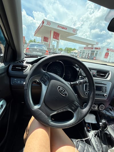 This media file shows a view from inside a car, looking out through the windshield. The car is at a Lukoil gas station, indicated by the prominent "ЛУКОЙЛ" (Lukoil) signage on the canopy and the red and white branding. The gas station also features a "МИНИМАРКЕТ" (Minimarket) sign.
The scene appears to be a sunny day with a sky full of white, fluffy clouds. Outside the car, several other vehicles are visible, suggesting a moderate amount of traffic or activity at the gas station. One car directly in front has the license plate "H962УС 193".
The interior of the car is also visible, including the steering wheel with the Kia logo, the dashboard with the speedometer, and the gear shift. A person's leg is visible on the right side of the frame, bathed in sunlight. The overall impression is a snapshot of a routine stop at a gas station on a bright day.