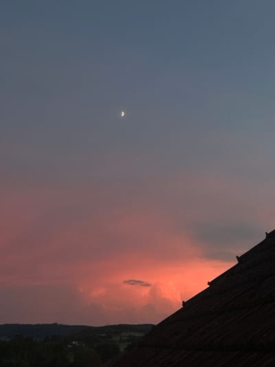 The image depicts a twilight sky with a crescent moon visible in the upper portion. The sky transitions from a deep blue at the top to vibrant shades of pink and orange closer to the horizon, indicating a sunset. Clouds are scattered throughout the sky, some illuminated by the setting sun. In the foreground, the dark silhouette of a building roof dominates the lower right section of the image. Beyond the roof, a distant landscape of rolling hills and trees is visible, suggesting a rural or semi-rural setting. The image was taken in Heinebach, Germany.