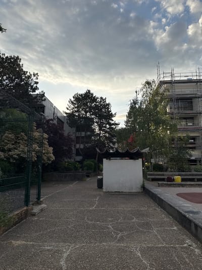 The photograph depicts an urban scene in Les Ulis, France. A multi-story building under renovation, scaffolding visible on its upper floors, is situated behind trees. A paved pathway, cracked with age, runs through the scene. A white tiled structure is positioned along the path. A metal fence borders a garden area filled with trees and flowering bushes. The sky is overcast with cloudy conditions. No people are visible in the shot.