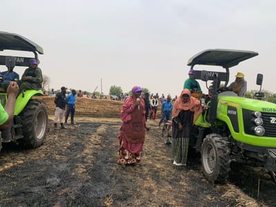 The image shows a scene in Ringimawa, Nigeria, a rural community located at the coordinates 11.666 latitude and 8.414 longitude. The setting is a field, likely a farm, with the ground covered in burnt grass and some remaining stalks. There are two green tractors in the foreground, one with the word "FAN" visible and the other with "WOFAN" on its side. People, mostly women, are gathered around the tractors. Several women are wearing traditional Islamic headwear. One woman, wearing a pink and black dress and a purple hat, is speaking, possibly addressing the group. Others are listening attentively. The overall atmosphere is one of community engagement and a possible gathering for a agricultural event or program.