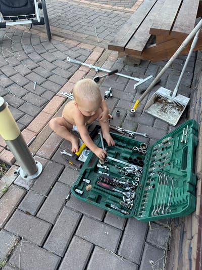 A Caucasian toddler, wearing only a diaper, sits on a paved patio area, looking down at an open green toolbox filled with various wrenches and sockets. The toolbox is positioned to the toddler's right. Scattered around the toddler and the toolbox are several individual tools, including wrenches and a socket attachment. In the background, there is a wooden picnic table and a broom with a dustpan. The patio is constructed of interlocking brick pavers, and there are patches of dry grass growing between the bricks. A metal post with a yellow top is visible to the toddler's left. The scene appears to be outdoors, likely in a residential yard. The location is identified as Борисьево Днп, Russia.