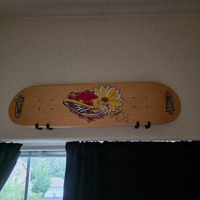 This media file shows a wooden skateboard mounted vertically on a textured, off-white wall. The skateboard has a graphic design featuring a yellow flower with red and white accents, and a red figure in a surfing pose with flames and a yellow surfboard with a blue logo. The skateboard also has two dark clips attached to it, presumably for mounting. There is a signature on the right side of the skateboard, above the graphic, with the text "Jeffrey Wet" and some other illegible writing. The top and bottom of the skateboard also feature a blue logo in an oval shape.
To the right of the skateboard, a black curtain is partially drawn, revealing a window with a view of trees and buildings outside. The sunlight streaming through the window suggests it is daytime. The setting appears to be an indoor space, possibly a bedroom or living area, in Beaverton, United States of America. The overall mood is casual and displays a piece of skateboarding memorabilia.