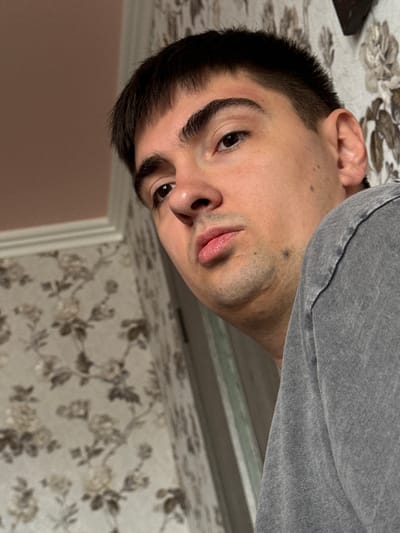 A close-up, low-angle, over-the-shoulder shot captures a young man with short, dark hair and dark eyes looking down and to the left. He has prominent eyebrows and a light stubble. His lips are slightly parted, and he is wearing a grey t-shirt. The background features floral wallpaper with a pattern of light-colored flowers and darker leaves against a light background. To the left, a wall painted a muted pink is visible, separated by a white trim. A portion of what appears to be a curtain or fabric with vertical stripes is also present. The overall lighting is dim. The image is set in a residential interior, likely a bedroom or living area, in the city of Kandry, Russia.