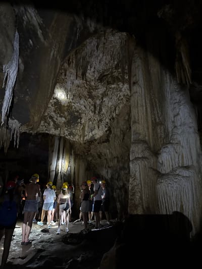 A group of tourists, all wearing hard hats, are gathered inside a dark cave. Stalactites and stalagmites are visible on the cave walls and ceiling, illuminated by the tourists' headlamps. The cave floor is uneven and appears to be made of rock and mud. The tourists are looking around the cave, some pointing at formations. The location is identified as Khao Phang, Thailand.