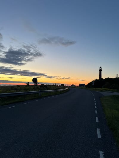 The image depicts a coastal road near Paldiski, Estonia, at dusk. The sky is a gradient of soft blues and oranges, with scattered clouds. A prominent red lighthouse stands silhouetted against the horizon on the right side of the frame, surrounded by trees. On the left, a road guardrail runs along the edge of the asphalt road, which curves away into the distance. A dark circular traffic sign is visible on the left side of the road. In the background, several small houses are clustered. The overall scene is peaceful and serene, capturing the transition from day to night.