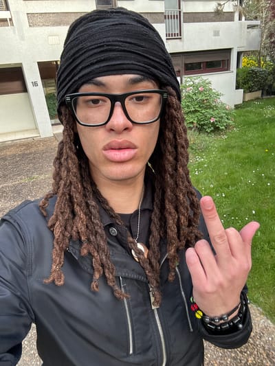 A person with brown dreadlocks, wearing a black head covering, black-framed glasses, and a black jacket is standing outdoors. They are giving the middle finger with their right hand, which also has a black beaded bracelet. The background includes a building labeled "16", grass, and shrubbery. The location appears to be in an urban area within Les Ulis, France.