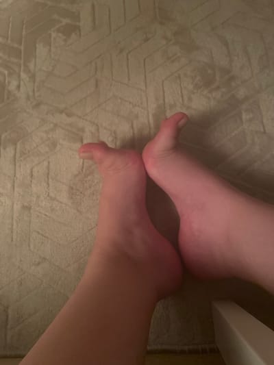 Two bare feet are shown from a top-down perspective, resting on a textured rug with a geometric pattern in a muted beige color. The feet are close together, with the toes of both feet pointing upwards and slightly to the left. The skin tone of the feet appears fair with a pinkish hue. The left foot is positioned slightly closer to the viewer and is curled upwards, with the toes clearly visible. The right foot is slightly behind and to the right of the left foot, and its sole is more prominently displayed. A sliver of a white object, possibly furniture, is visible in the bottom right corner of the image. The lighting appears to be dim, casting subtle shadows. The image is an indoor scene, and there is no indication of the specific location in Yunus Emre, Turkey, beyond it being an indoor setting.