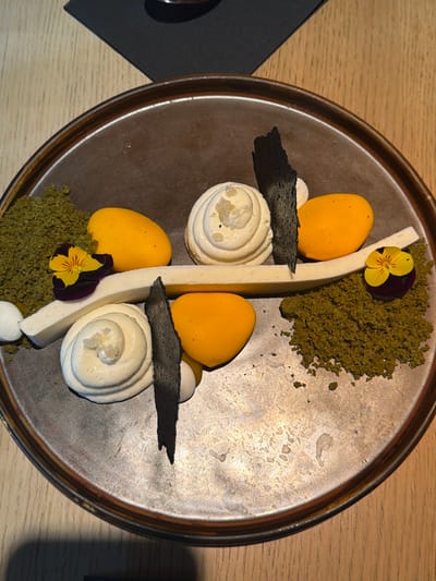 The scene depicts a meticulously arranged dessert on a round, textured metallic-bronze plate, situated on a light wooden table surface. In the upper left background, a dark grey fabric napkin partially covers a dark object. The dessert features two mounds of green, granular crumble; one larger on the left, one smaller on the right. A curved, off-white bar-shaped dessert element traverses the plate diagonally from lower-left to upper-right, with small white spherical components at its ends. Two distinct swirls of white cream, each topped with fine crystalline particles, are present centrally on the plate, one above and one below the diagonal bar. Four vibrant orange, ovoid-shaped pieces are distributed across the plate. Two irregular, dark charcoal-colored shards are positioned vertically within the arrangement. Two small edible flowers with purple and yellow petals are placed on the plate; one on the left adjacent to the larger green crumble and one on the right near the smaller green crumble. No individuals, actions, or direct interactions are visible. The location is Kiryat Ono, Israel.