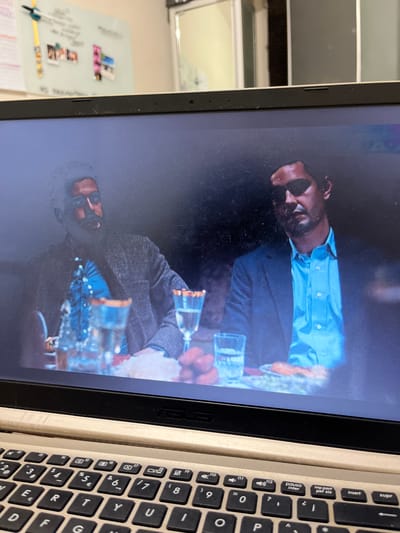 The image shows a laptop screen displaying a scene from a movie or show. Two men are seated at a table, with drinks and food in front of them. The man on the left is wearing a tweed jacket over a blue shirt, and the man on the right is wearing a suit jacket over a light blue shirt. The scene appears to be indoors, possibly a restaurant or a formal setting. The background on the screen is dark and indistinct. The laptop itself is open, and part of its keyboard is visible in the foreground. The setting is an indoor environment, with a whiteboard visible in the blurred background of the room the laptop is in. There is no indication in the image that this scene is located in San Luis, Peru.